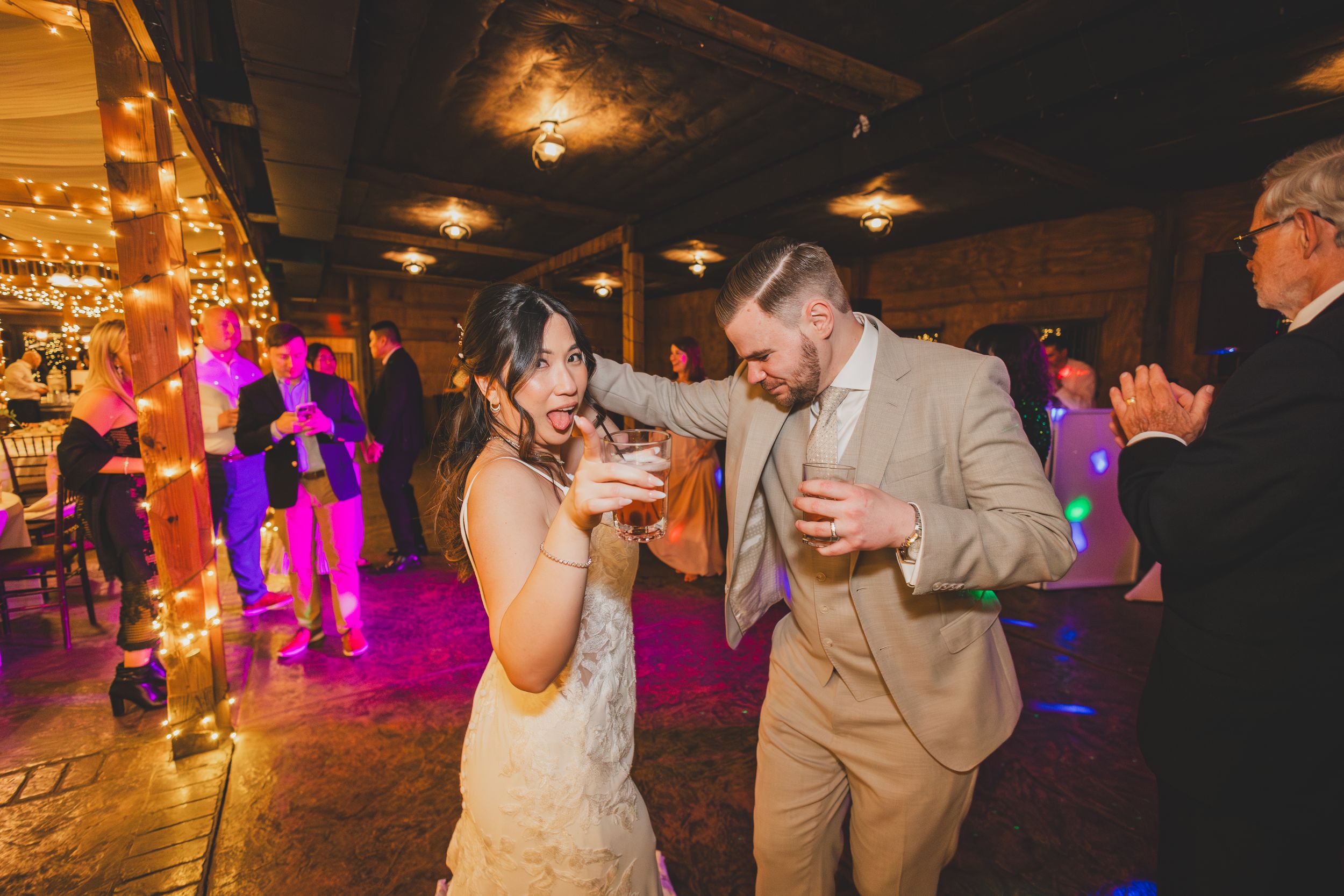 Wedding guests dancing and celebrating on a dimly lit dance floor with colorful purple lighting and warm ambient glow.