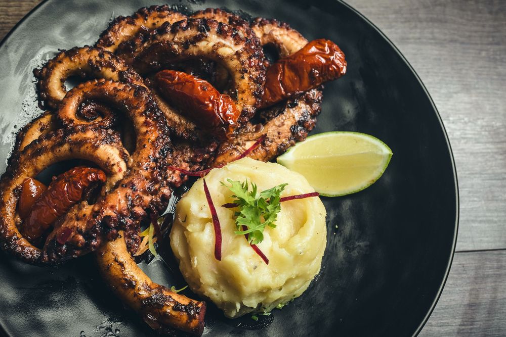 Grilled octopus served with mashed potatoes and lime on black plate.