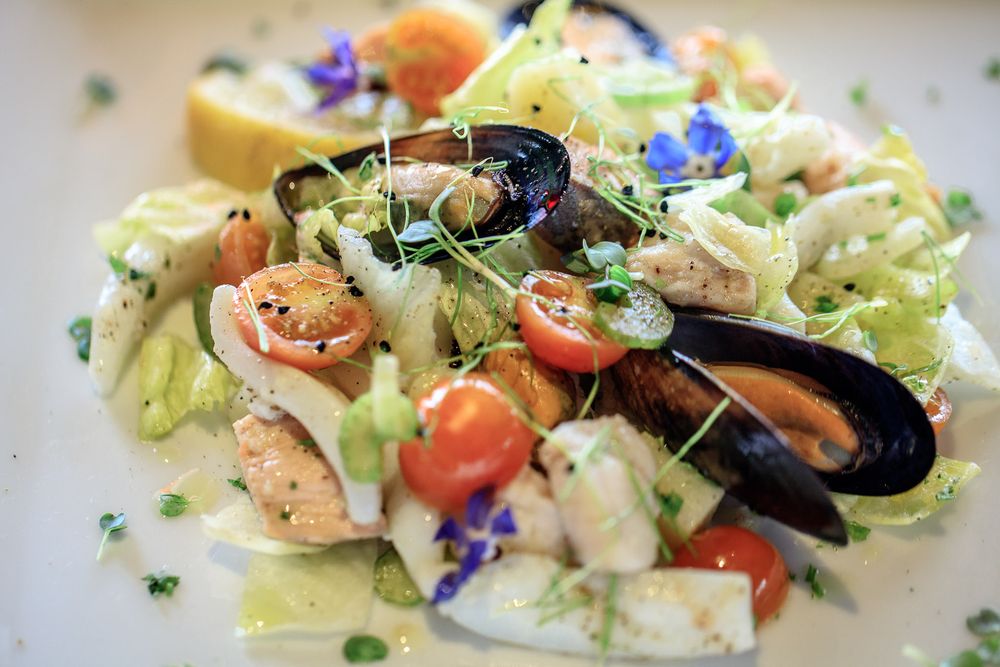 Seafood salad with mussels, cherry tomatoes and blue edible flowers on white plate.