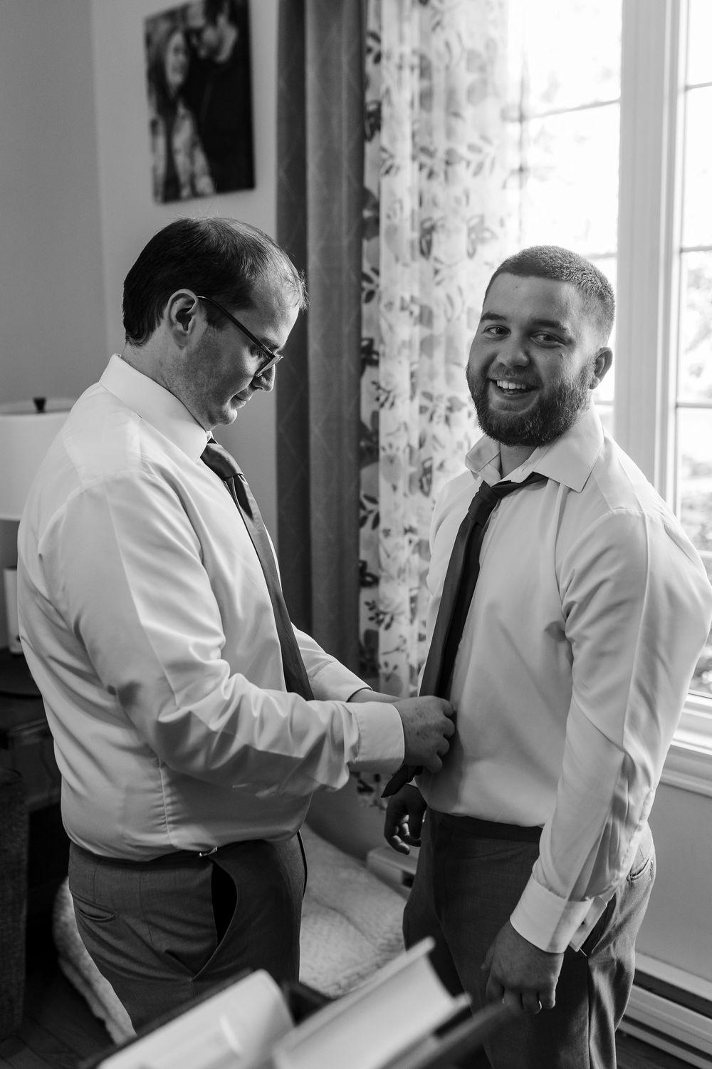 Two people in white dress shirts and ties getting ready in a room with sheer curtains and natural light.
