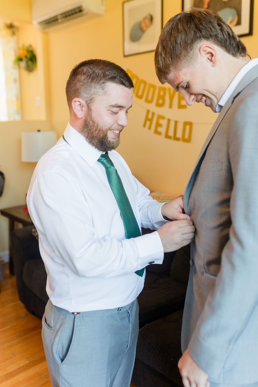 Two people in formal wear adjusting ties and helping each other prepare in a room with yellow walls.