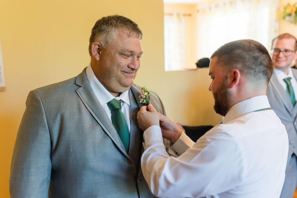 A person helps adjust a boutonniere on a wedding party member wearing a grey suit with green tie.