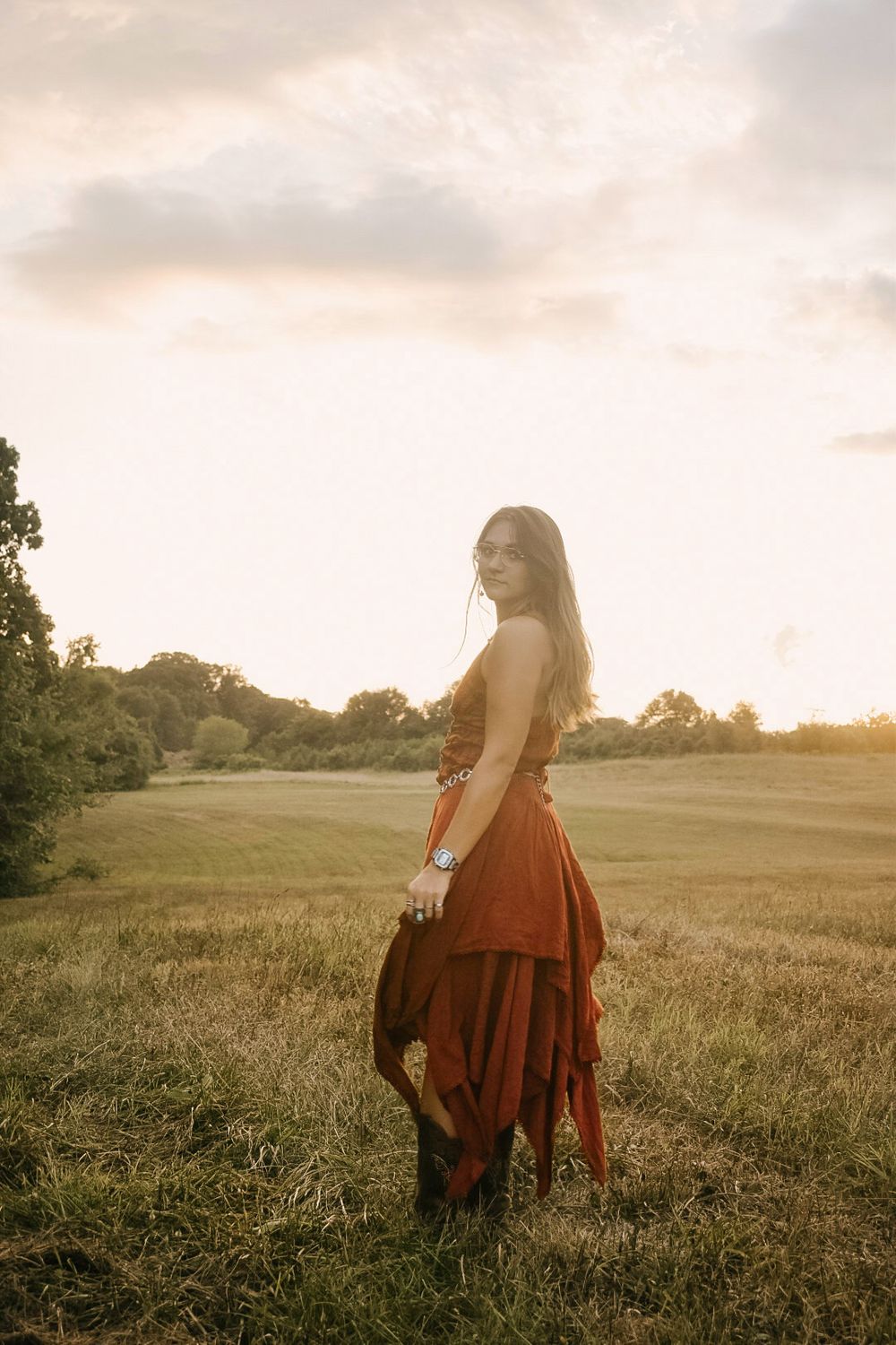 A figure in a flowing red dress stands in a meadow at golden hour with sunlight streaming through trees in the background.