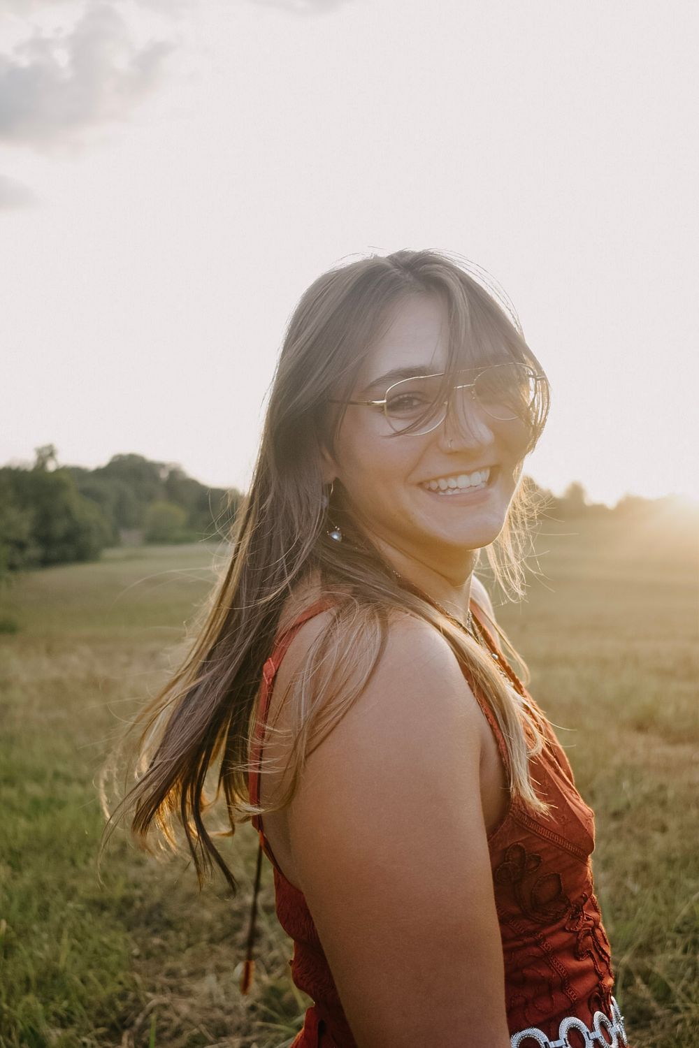 A figure in a red dress smiles warmly in a grassy field during golden hour sunset.