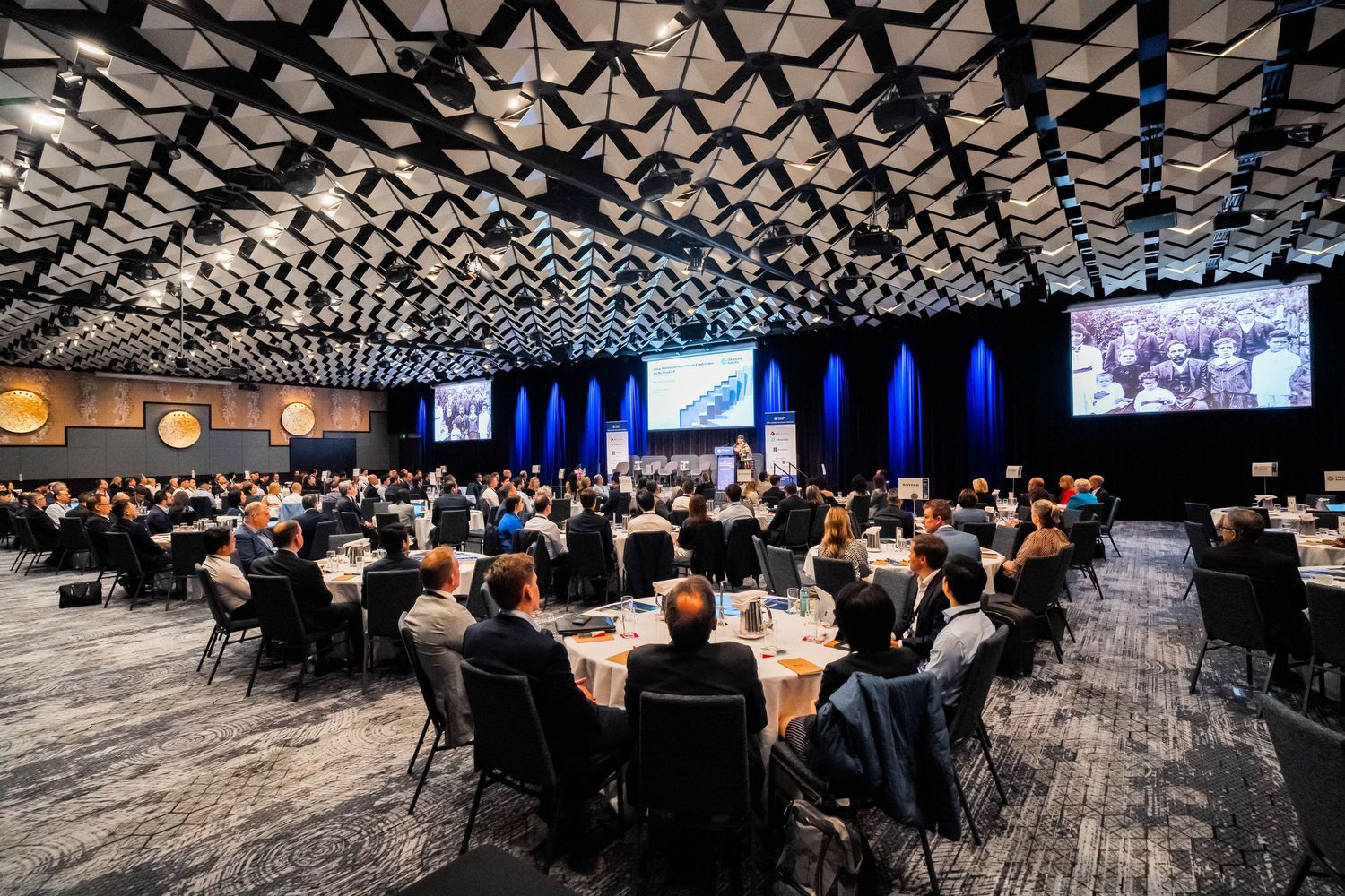 Business conference attendees seated in rows in a large modern auditorium with geometric patterned ceiling.