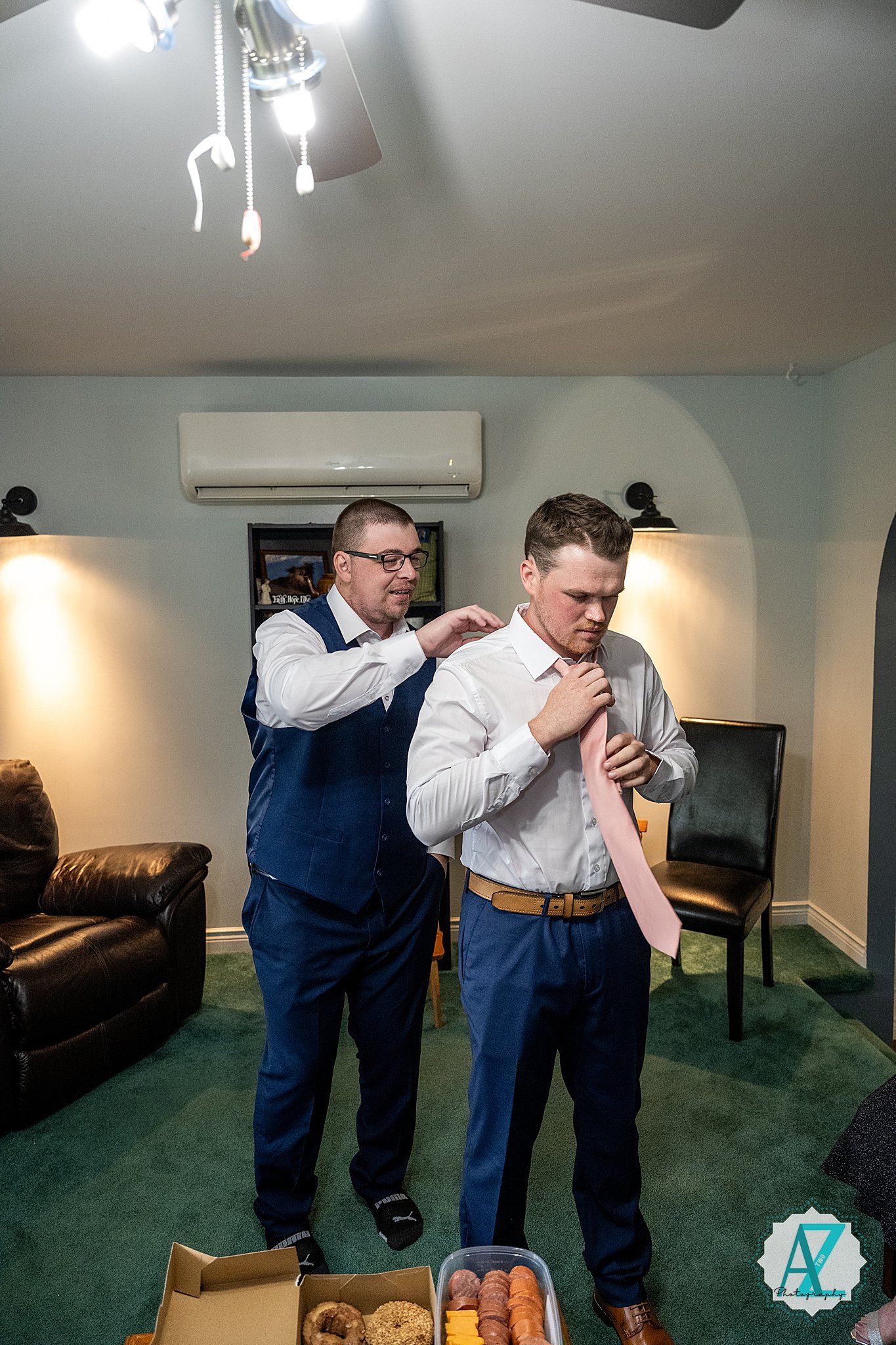 Two people in blue vests and white shirts help each other with formal attire preparations in a carpeted room.