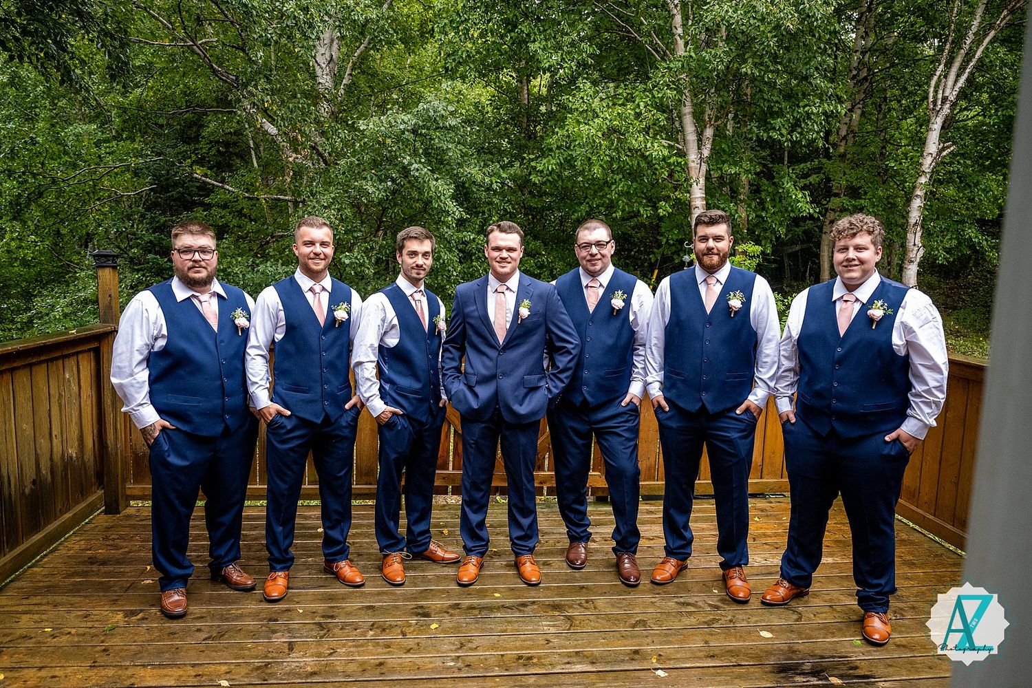 Groomsmen in navy vests and white shirts pose together on wooden deck surrounded by greenery.