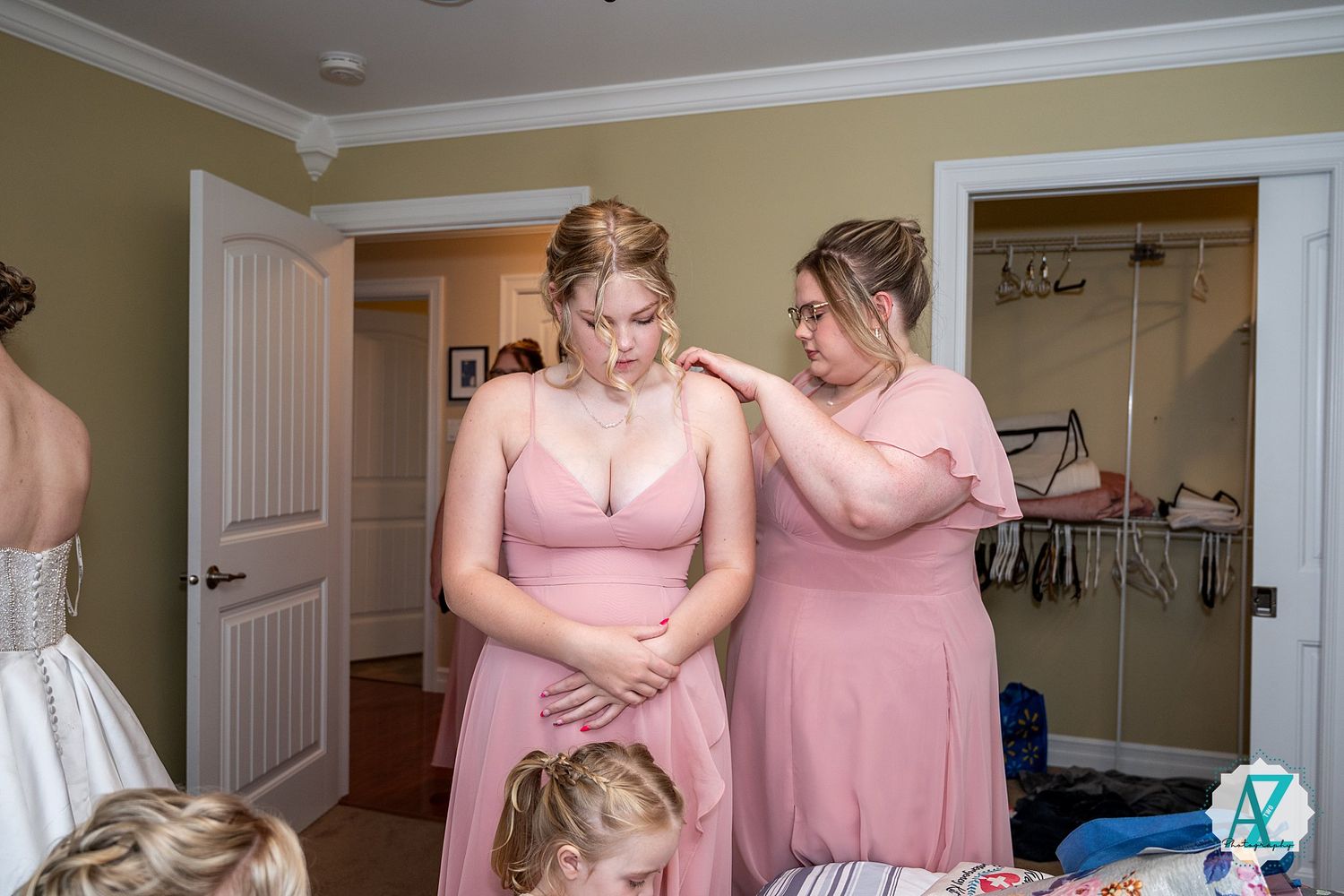 Bridesmaids wearing pink dresses help each other prepare in a cream-colored room before the wedding ceremony.