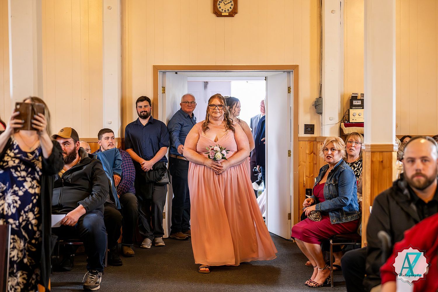A wedding party entrance procession in a chapel hallway with guests seated along the walls, captured in warm lighting.