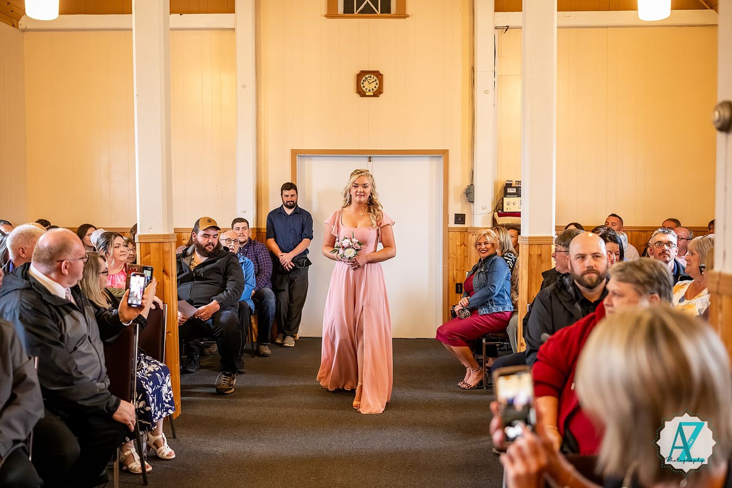 Wedding party member in pink gown walks down church aisle as seated guests watch during ceremony.