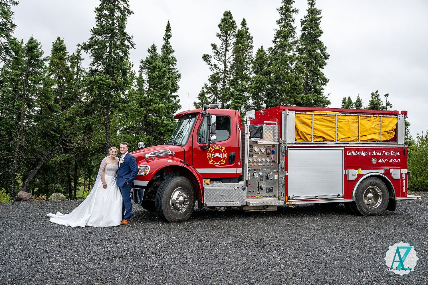 A couple takes wedding photos with a vintage red fire truck parked on a gravel road amid evergreen trees.