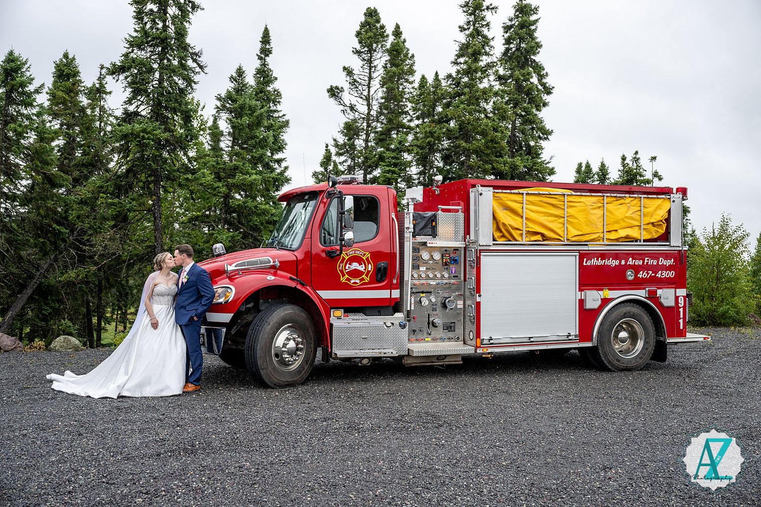 A red fire truck parked in a gravel lot surrounded by evergreen trees on a cloudy day.