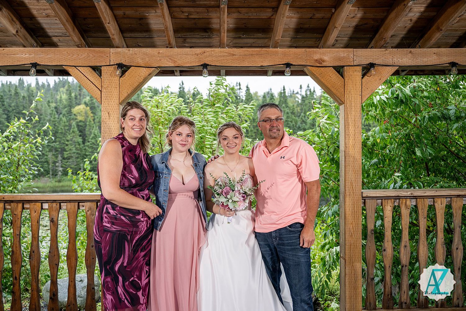 Celebratory group gathers under a wooden pavilion for outdoor wedding photos in summer garden setting.