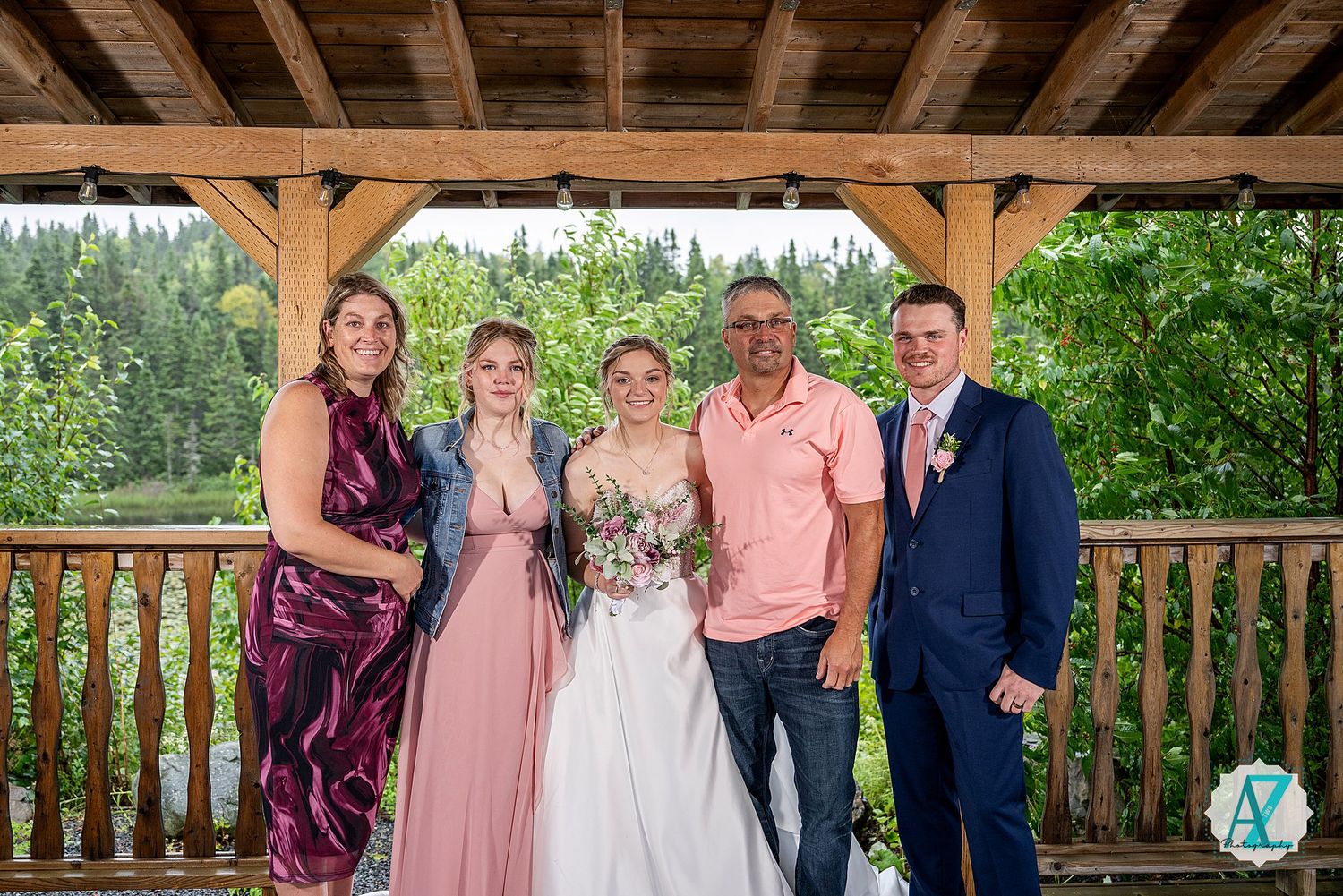Wedding party members pose together under a rustic wooden pavilion with outdoor scenery behind them.