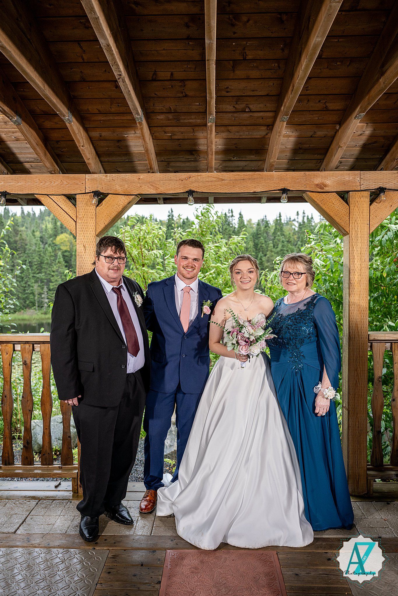 Wedding party gathered under a rustic wooden pavilion with exposed beams on a sunny outdoor day.