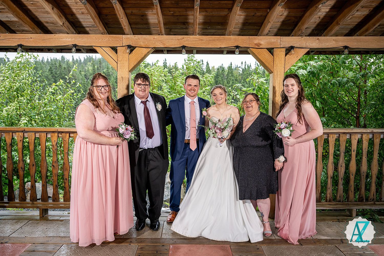 Wedding party and attendants in pink dresses and formal attire pose beneath a wooden gazebo structure.