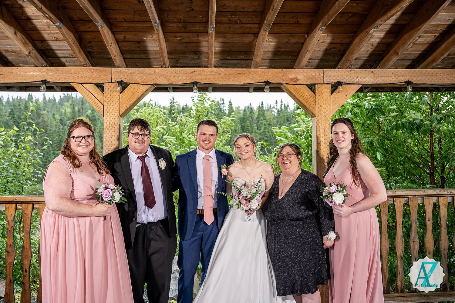 Wedding party poses in elegant pink and navy attire under rustic wooden pavilion surrounded by greenery.