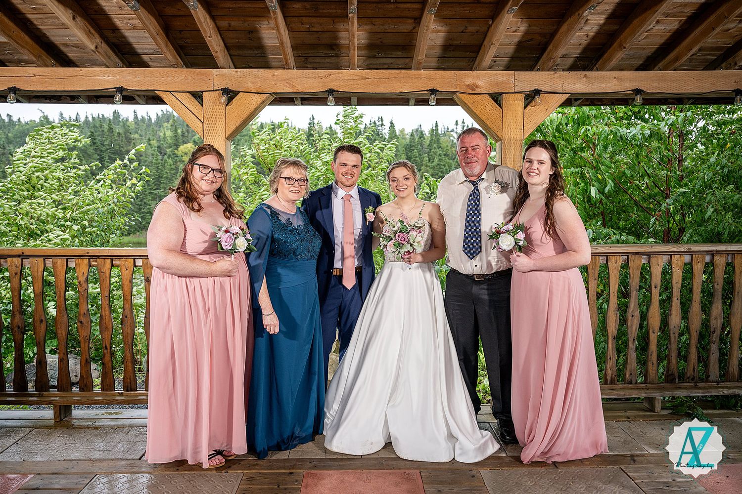 Group photo of bridal party wearing pink dresses and dark suits standing in an outdoor wooden pavilion.