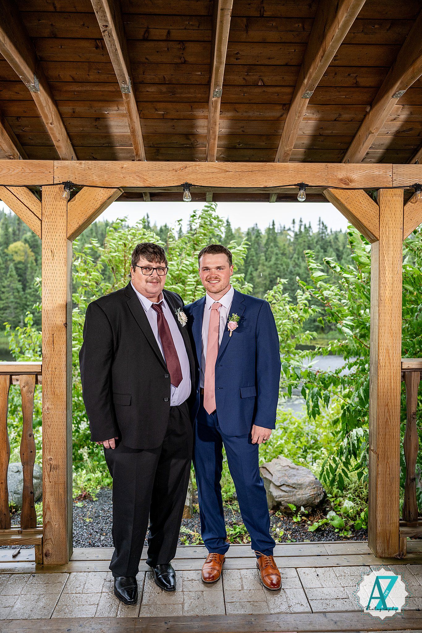 Groomsmen in navy and burgundy suits pose casually under wooden beams of outdoor pavilion.