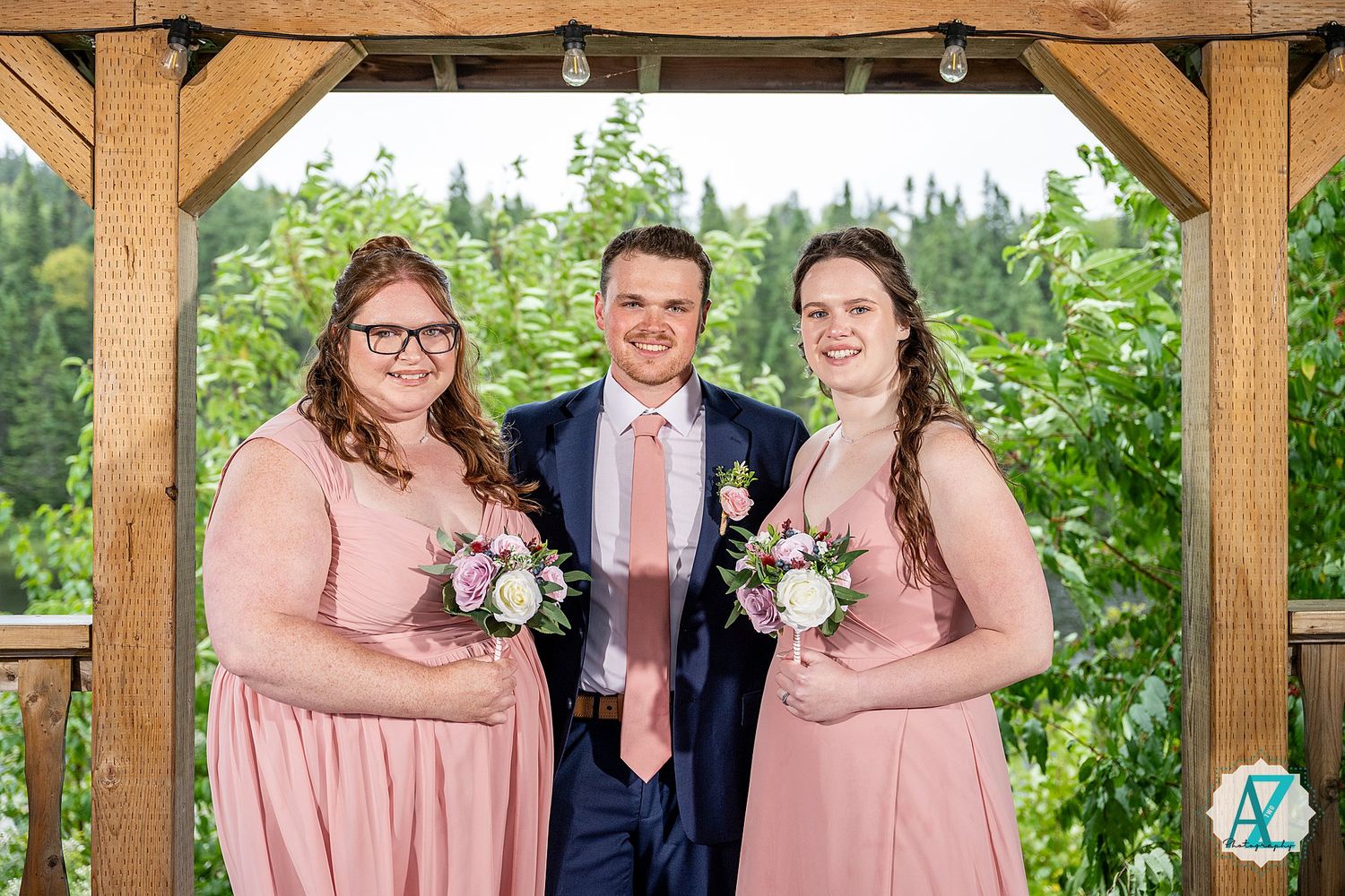 Wedding party poses in blush pink dresses and navy suits under a wooden pergola surrounded by greenery.