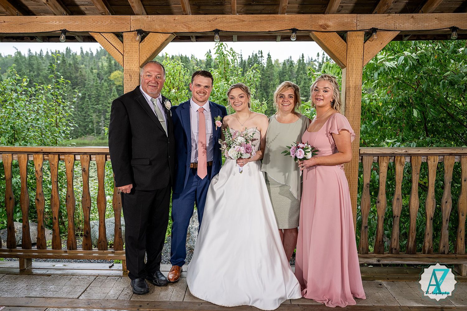 Wedding party members gather for formal photos under a covered wooden deck with scenic views.