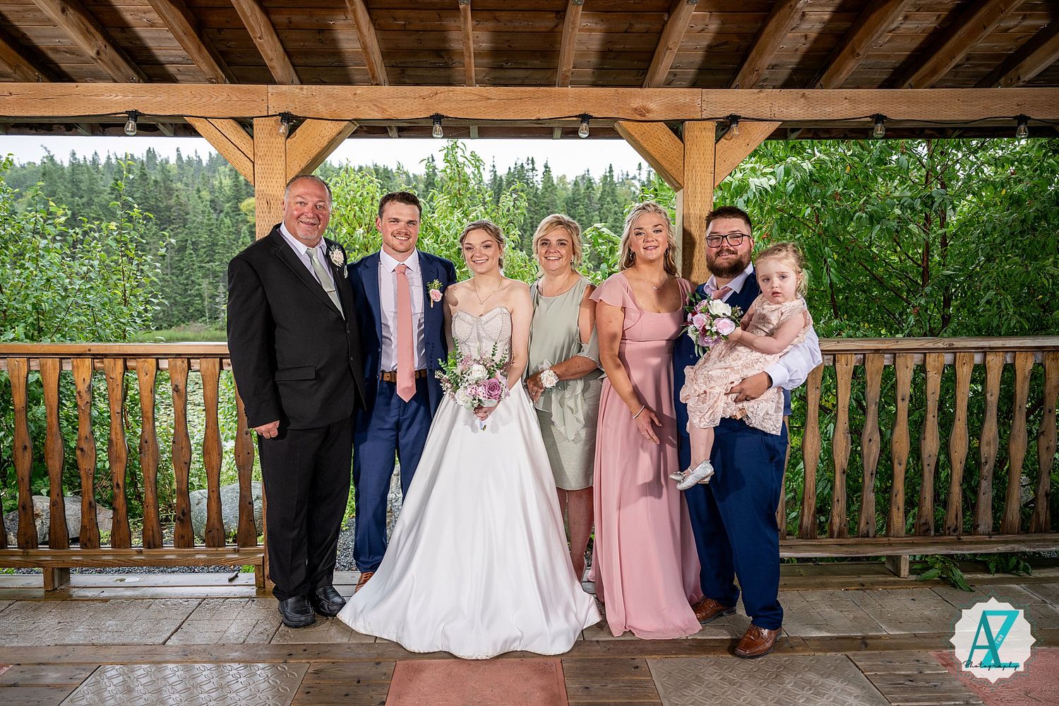 Wedding party poses on rustic wooden pavilion surrounded by lush green trees on a summer day.