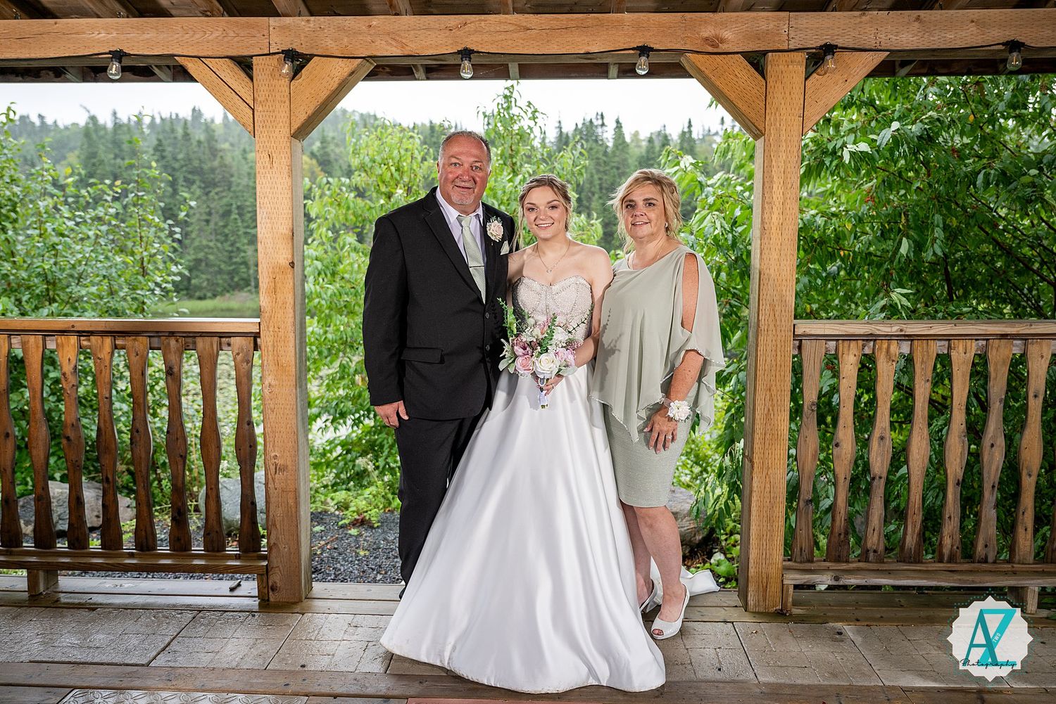Wedding party poses under a rustic wooden gazebo surrounded by lush green trees and forest scenery.