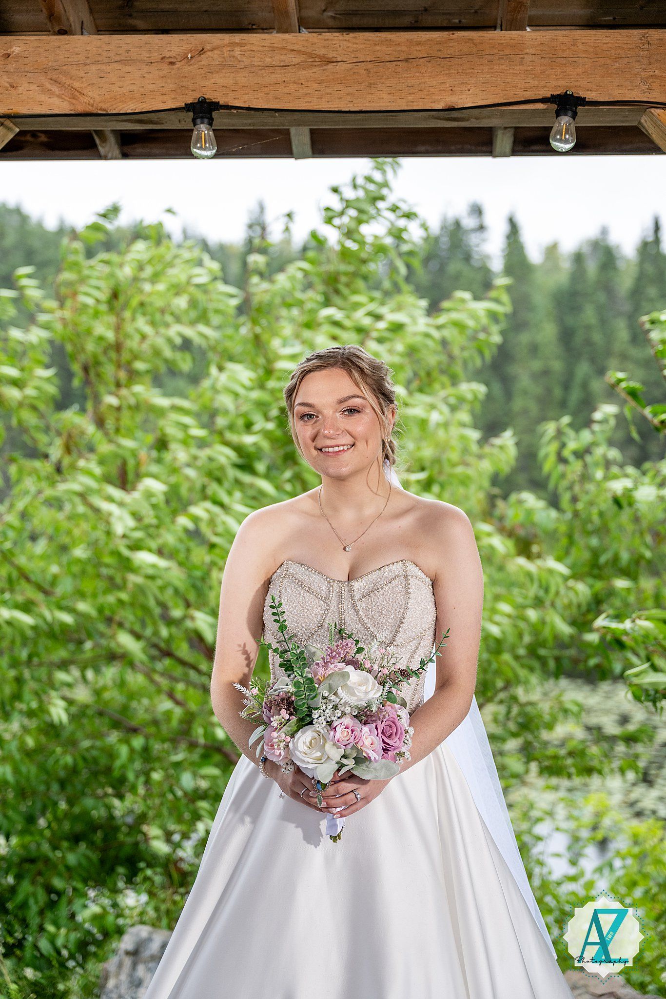A wedding portrait under a rustic wooden pergola with lush greenery and evergreen trees in the background.