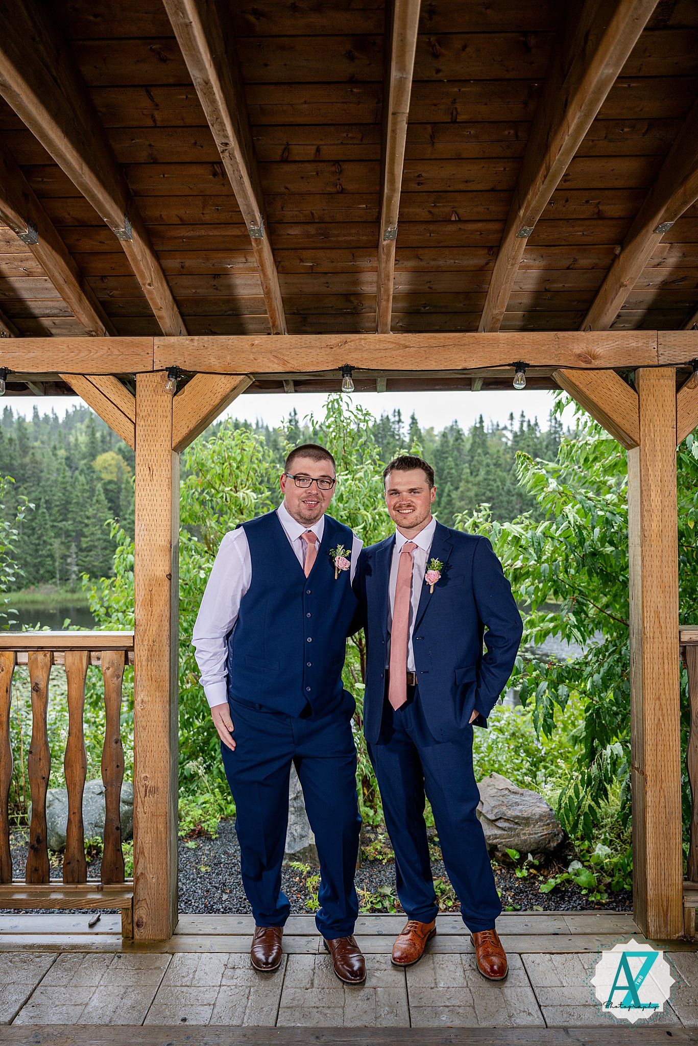 Three groomsmen in navy suits and coral ties sitting on rustic wooden porch with forest views.
