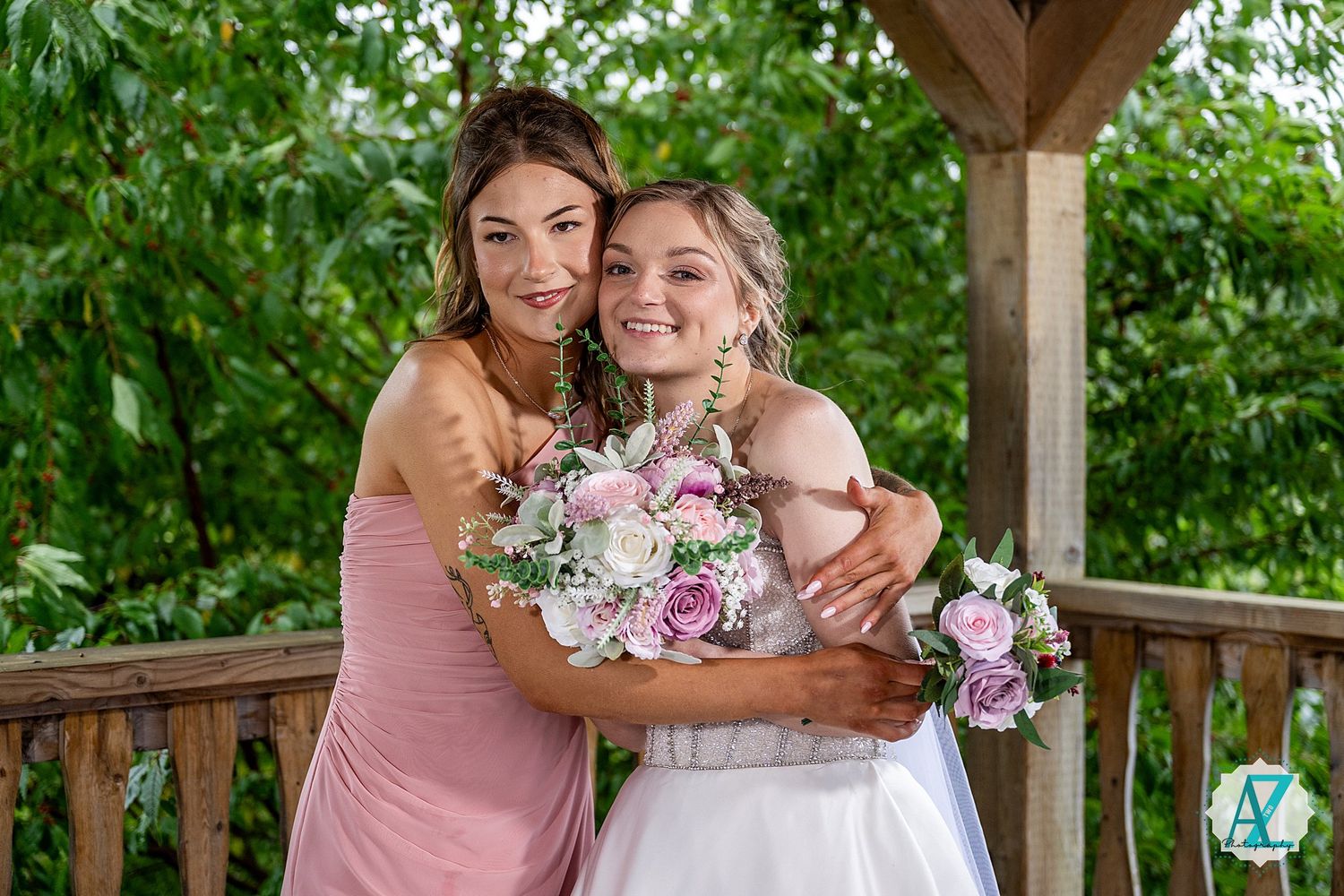 Wedding group poses on wooden deck against backdrop of vibrant greenery and summer foliage.
