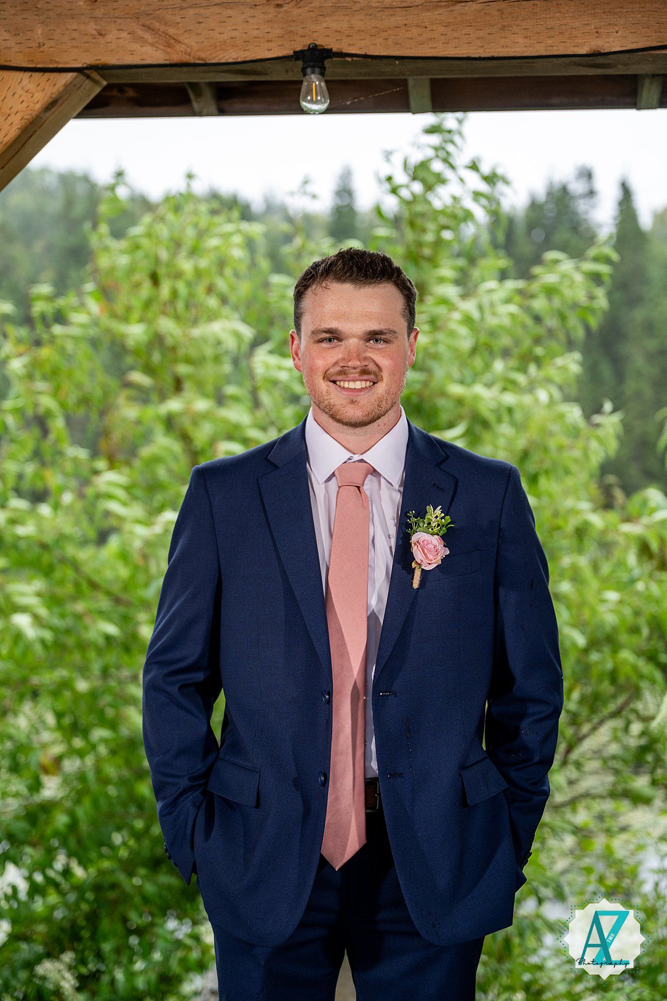 Groomsman in navy blue suit with peach tie poses outdoors against greenery backdrop.