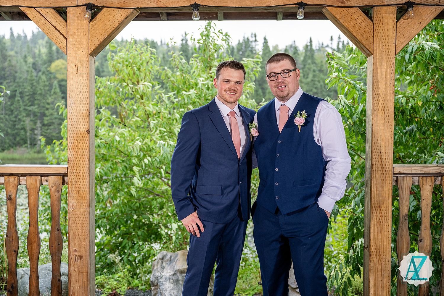 Group in matching navy vests and formal attire pose beneath a rustic wooden gazebo during an outdoor celebration.