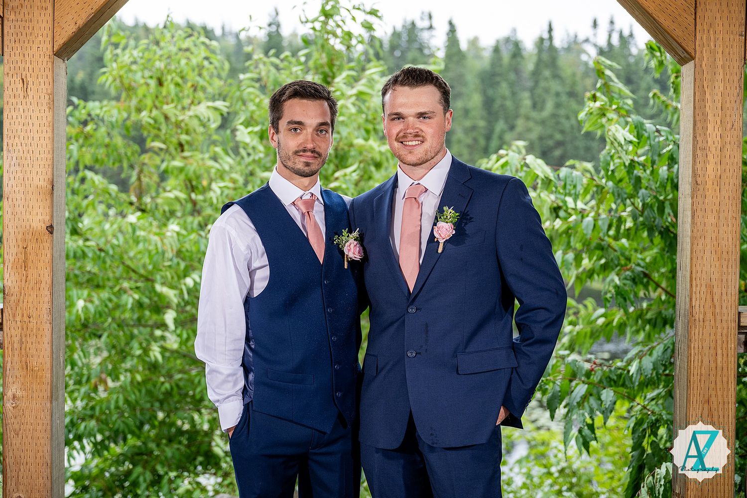 Two people wearing navy suits stand together under a wooden pergola surrounded by lush greenery.