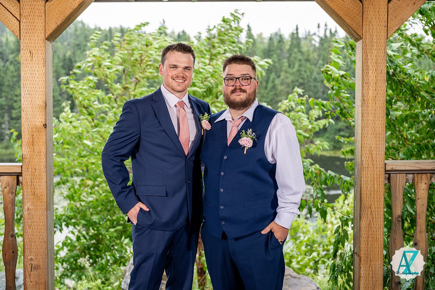 Two people wearing suits and ties stand together at an outdoor wooden pavilion surrounded by green foliage.