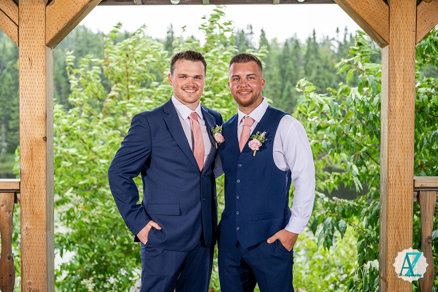 Two people wearing suits and ties stand together at an outdoor wooden pavilion surrounded by green foliage.