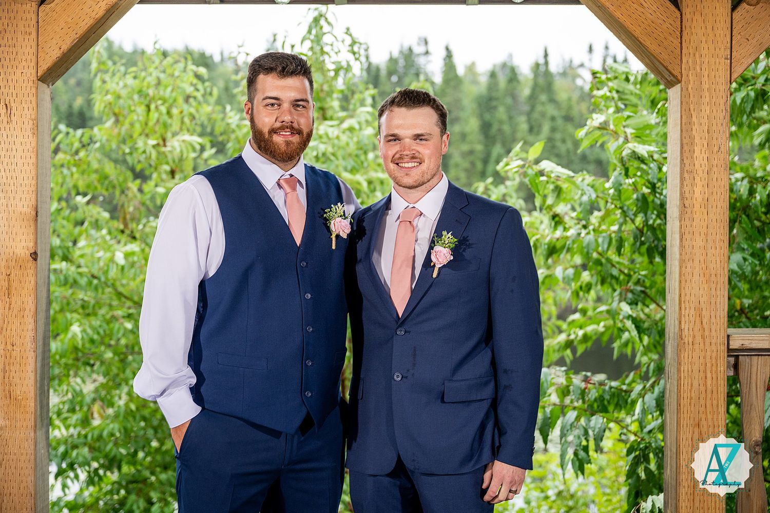 Two people in navy suits stand together smiling in a garden setting with a wooden structure overhead.