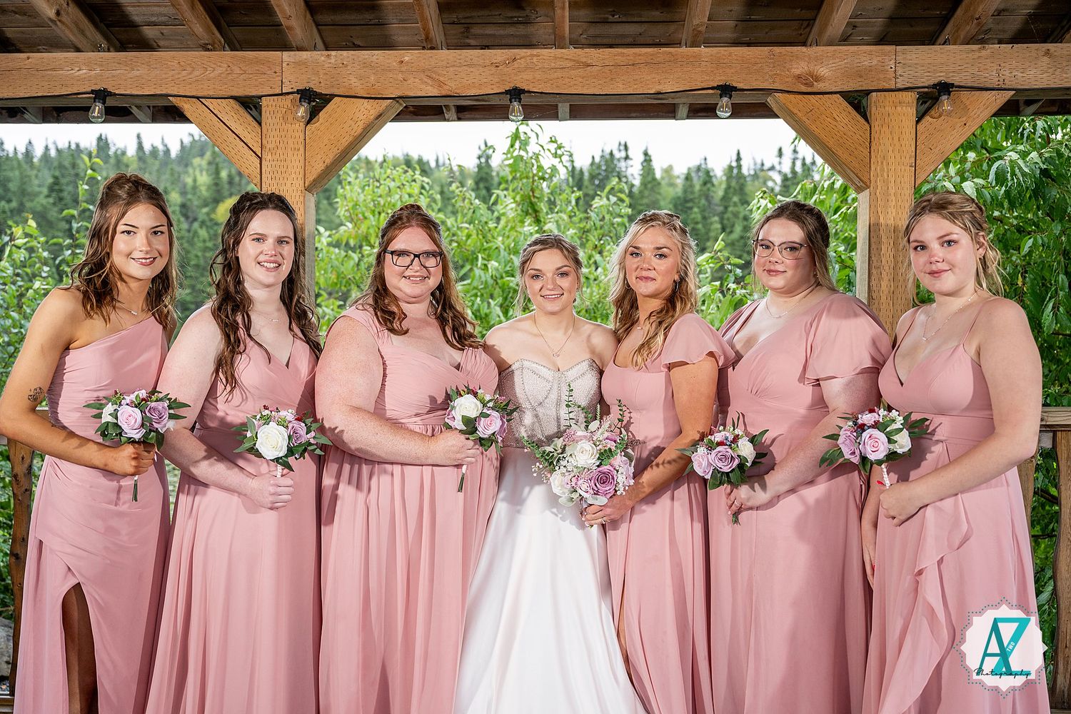 Wedding party in dusty rose bridesmaid dresses and white bridal gown pose together under wooden pavilion in outdoor setting.
