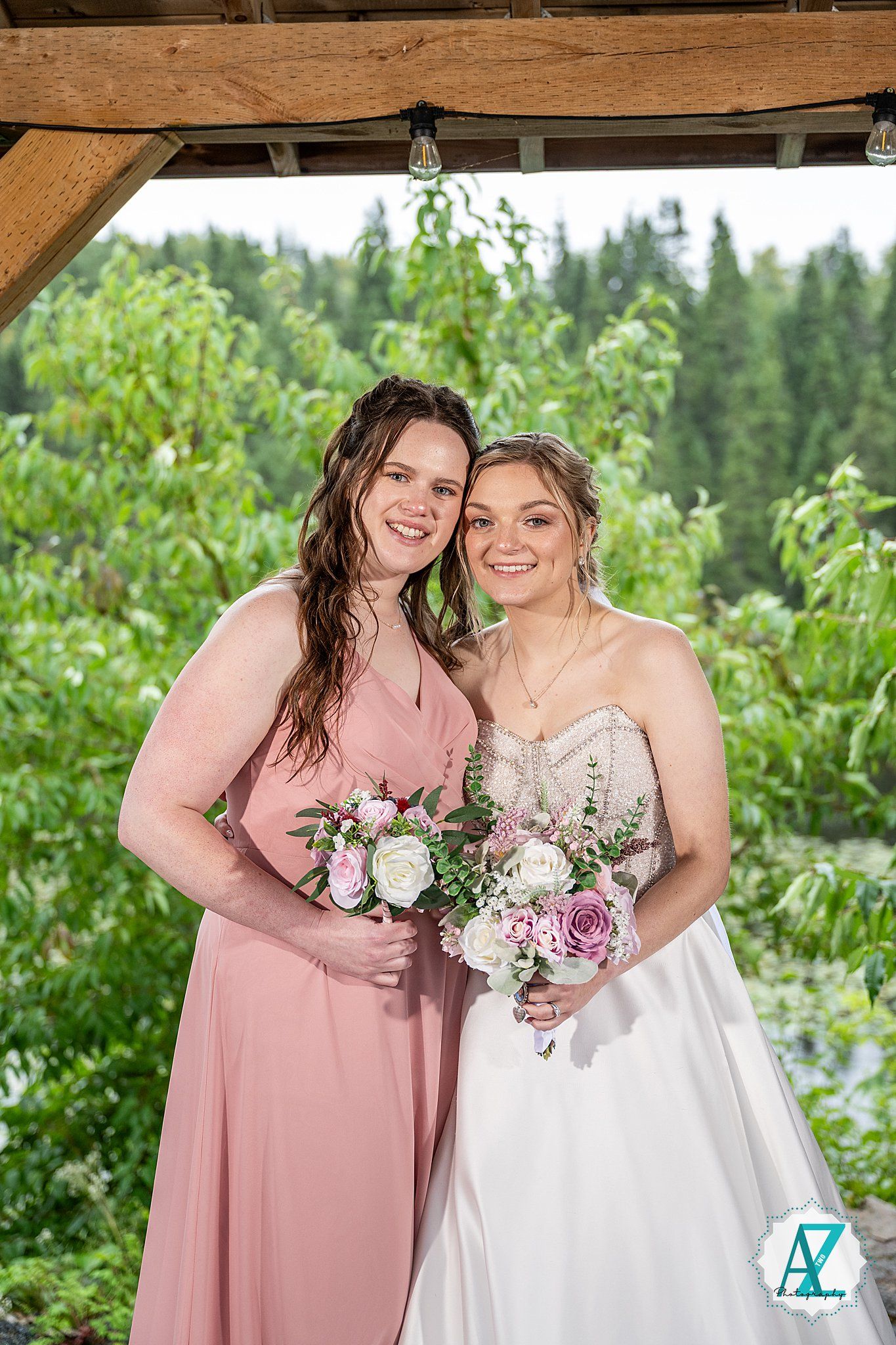 Wedding party standing outdoors in pink and white dresses holding floral bouquets against natural greenery.