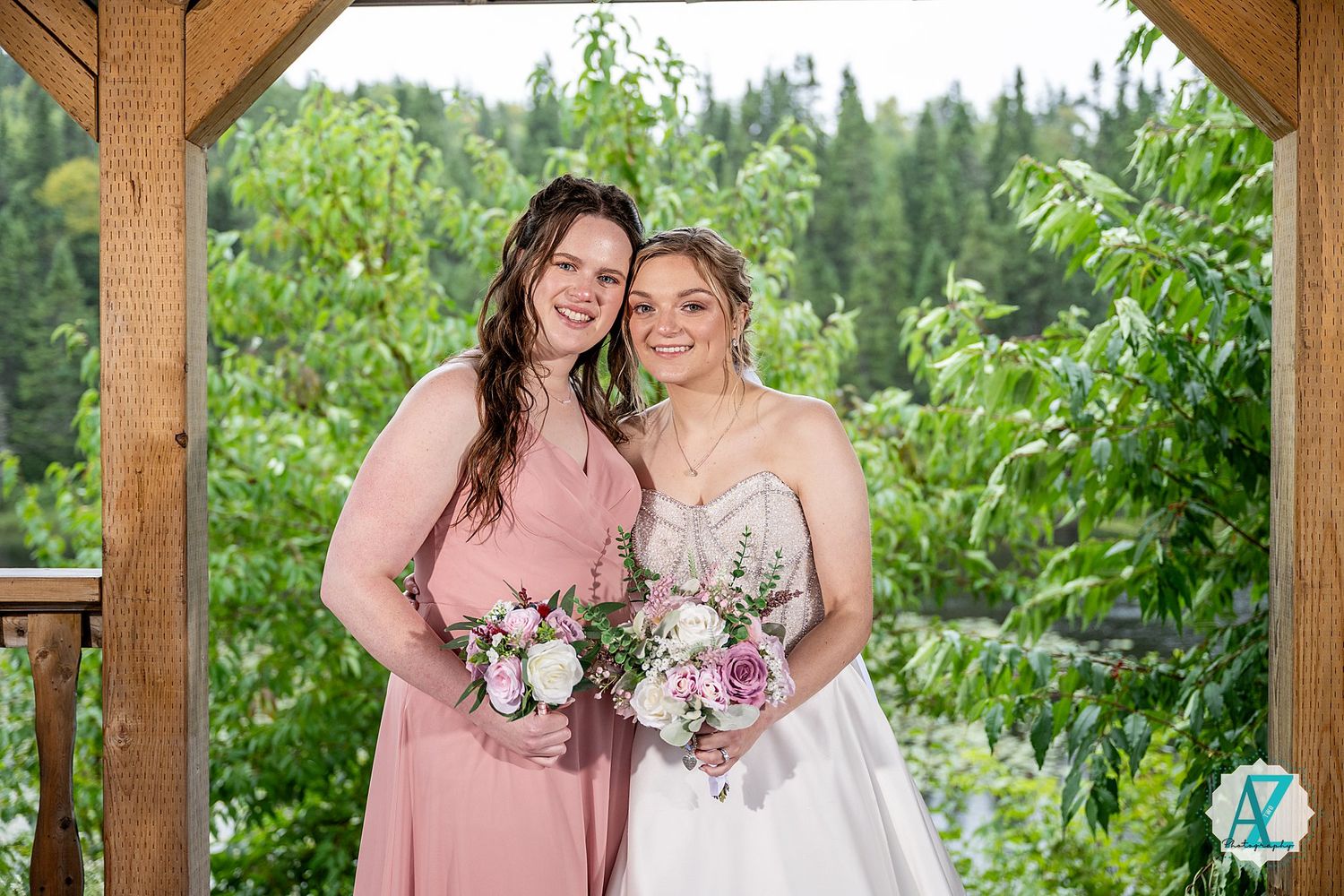 Two figures in pink and white dresses pose under a wooden gazebo surrounded by lush greenery holding floral bouquets.