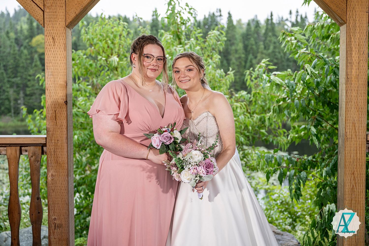 Two people stand together under a wooden gazebo holding pink and purple flower bouquets against a forest backdrop.