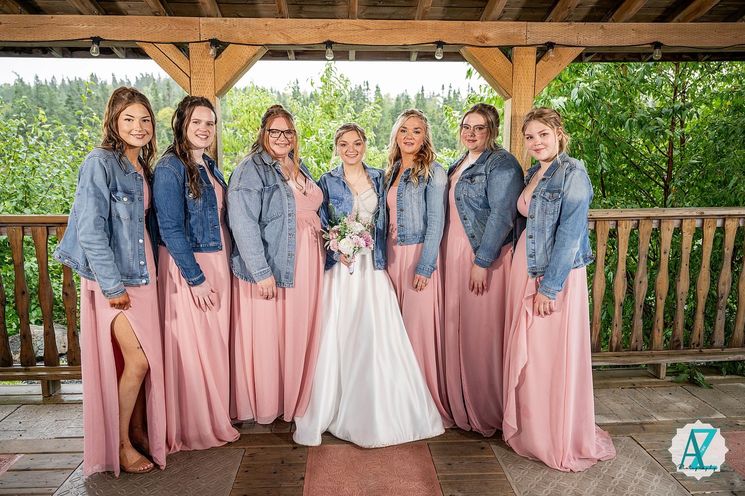 A line of bridesmaids in dusty rose dresses and denim jackets pose together at a rustic outdoor venue.
