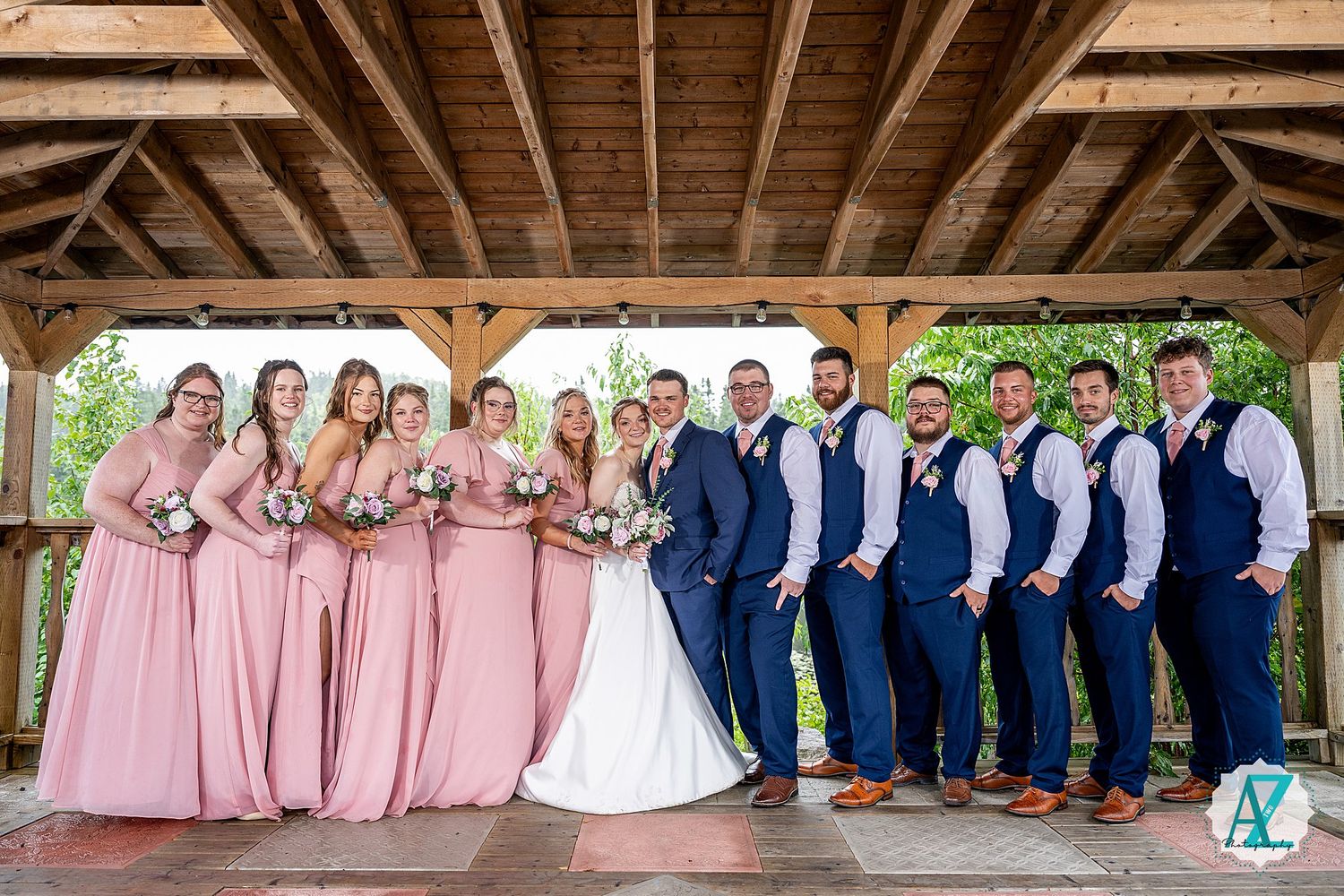 Wedding party in pink bridesmaid dresses and navy suits gather under a rustic wooden pavilion for group photos.