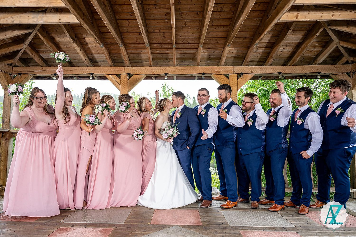 Wedding party members in pink dresses and navy suits line up under a rustic wooden pavilion.