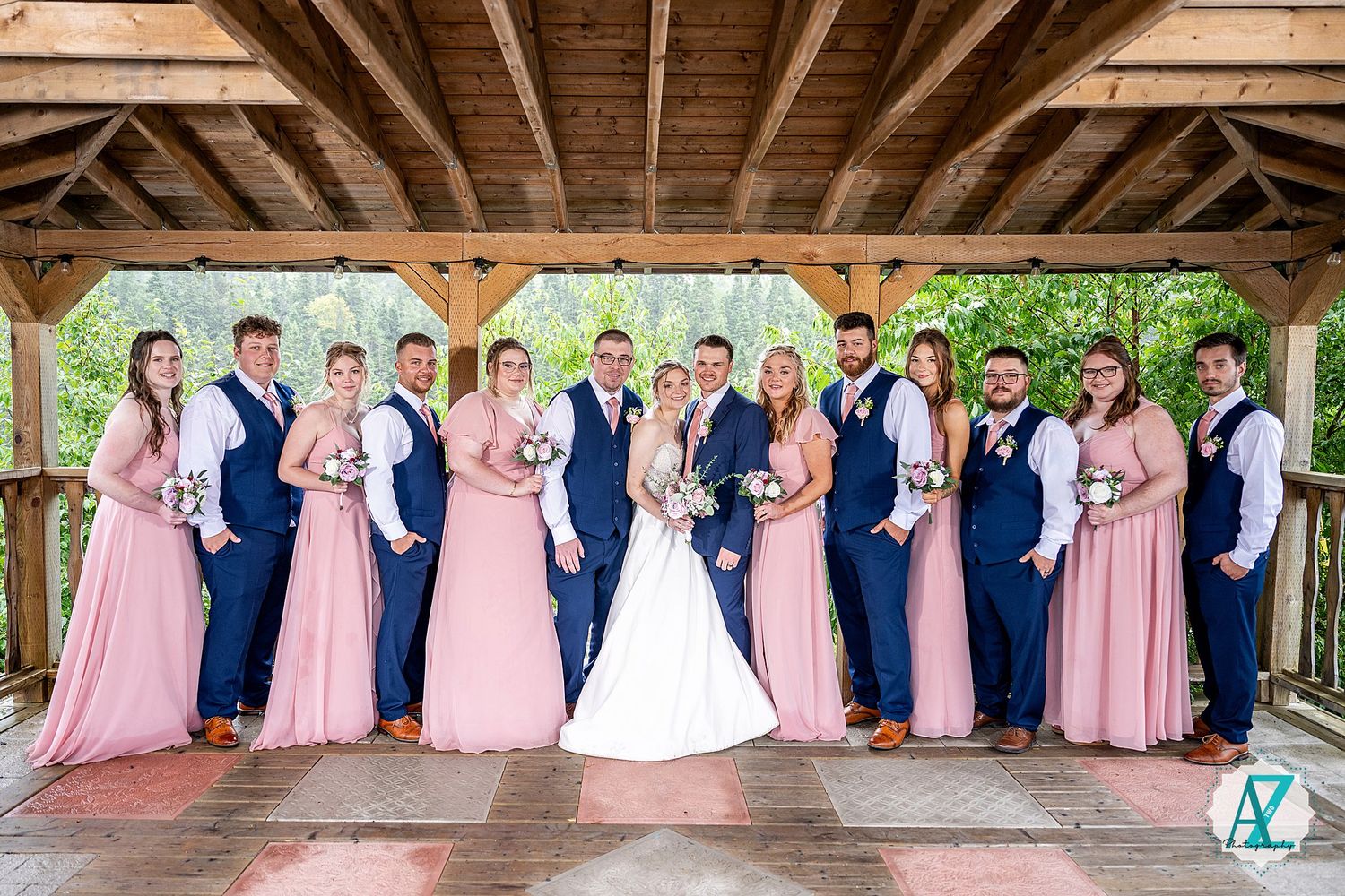 A wedding party in navy blue and pink attire poses together under a rustic wooden pavilion structure.
