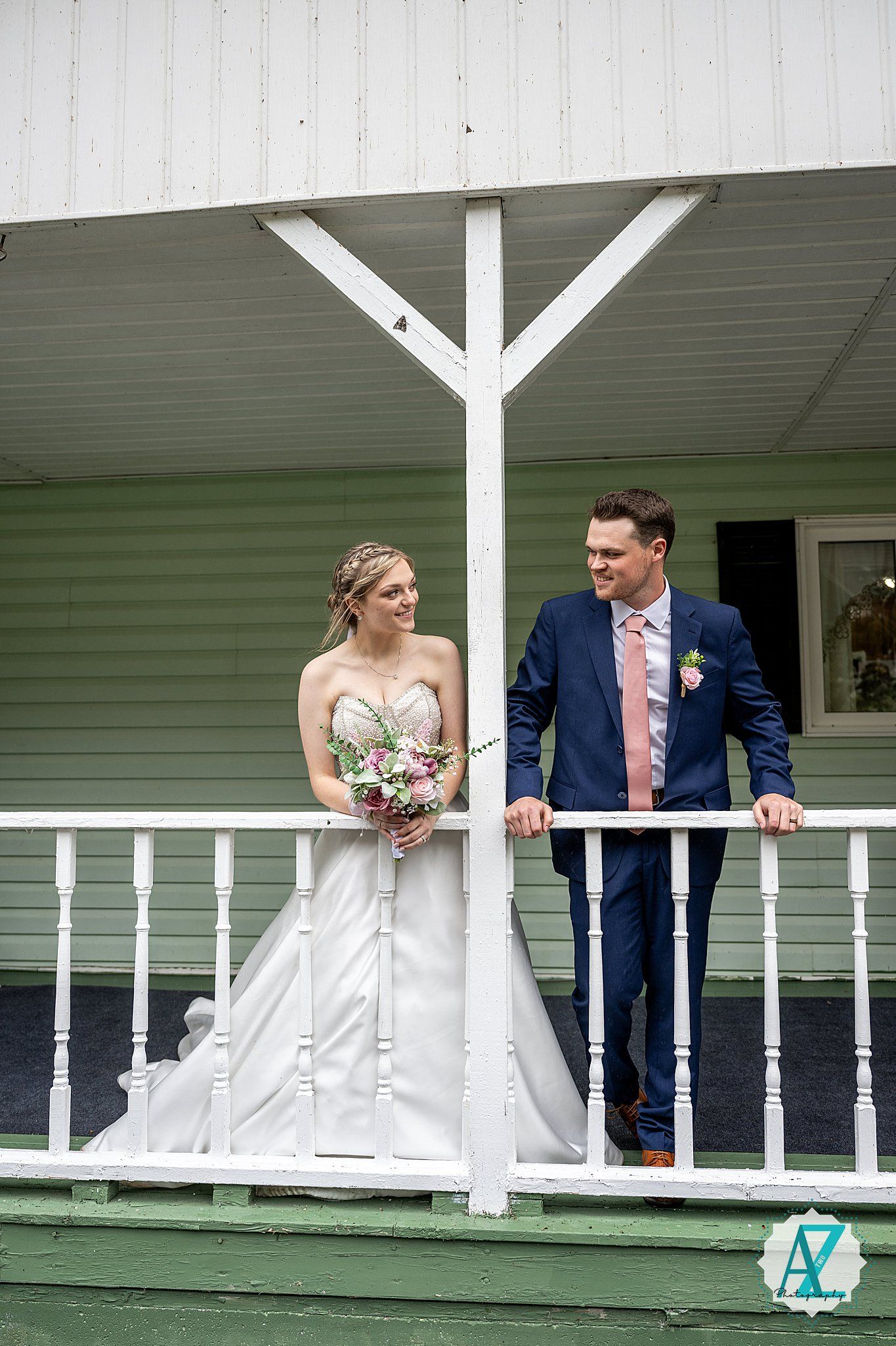 A wedding couple poses playfully along a white railing on a green porch at their outdoor venue.
