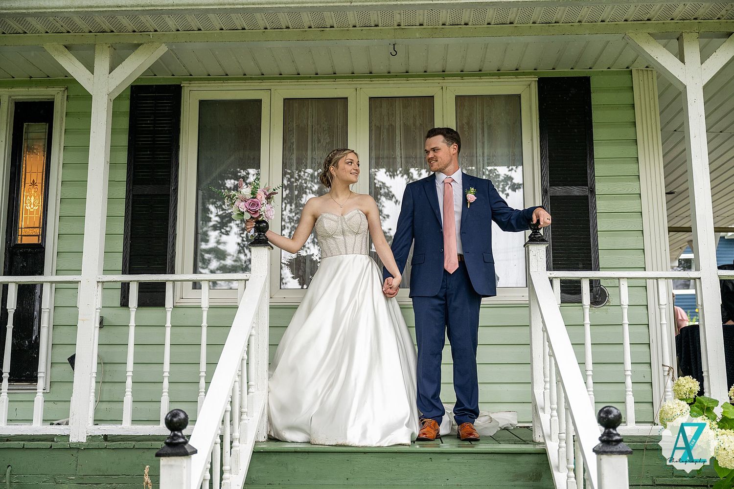A couple stands together on a classic white porch of a mint green house during a wedding celebration.