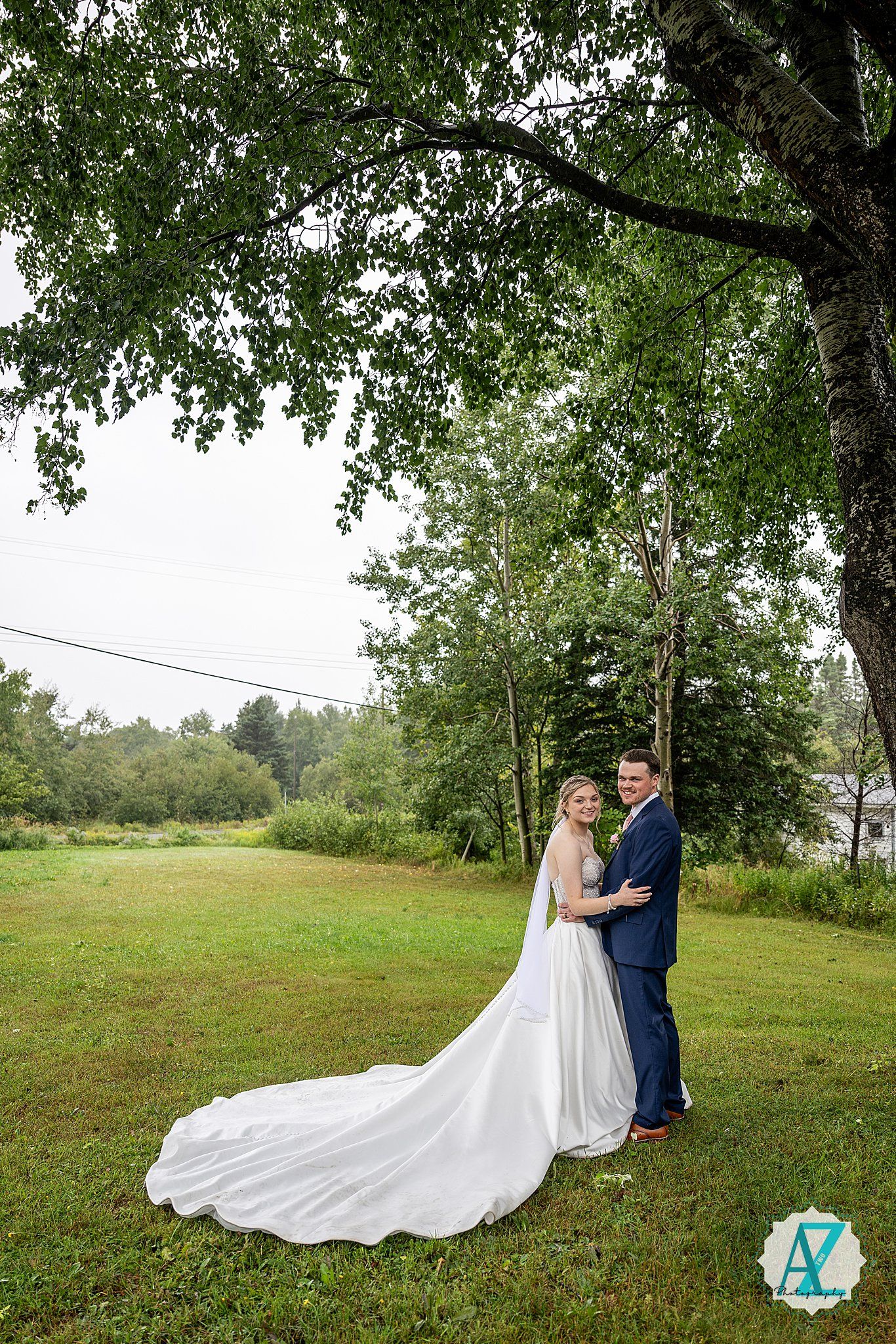 Wedding couple stands in tranquil outdoor setting with flowing white dress trailing across grassy hillside.