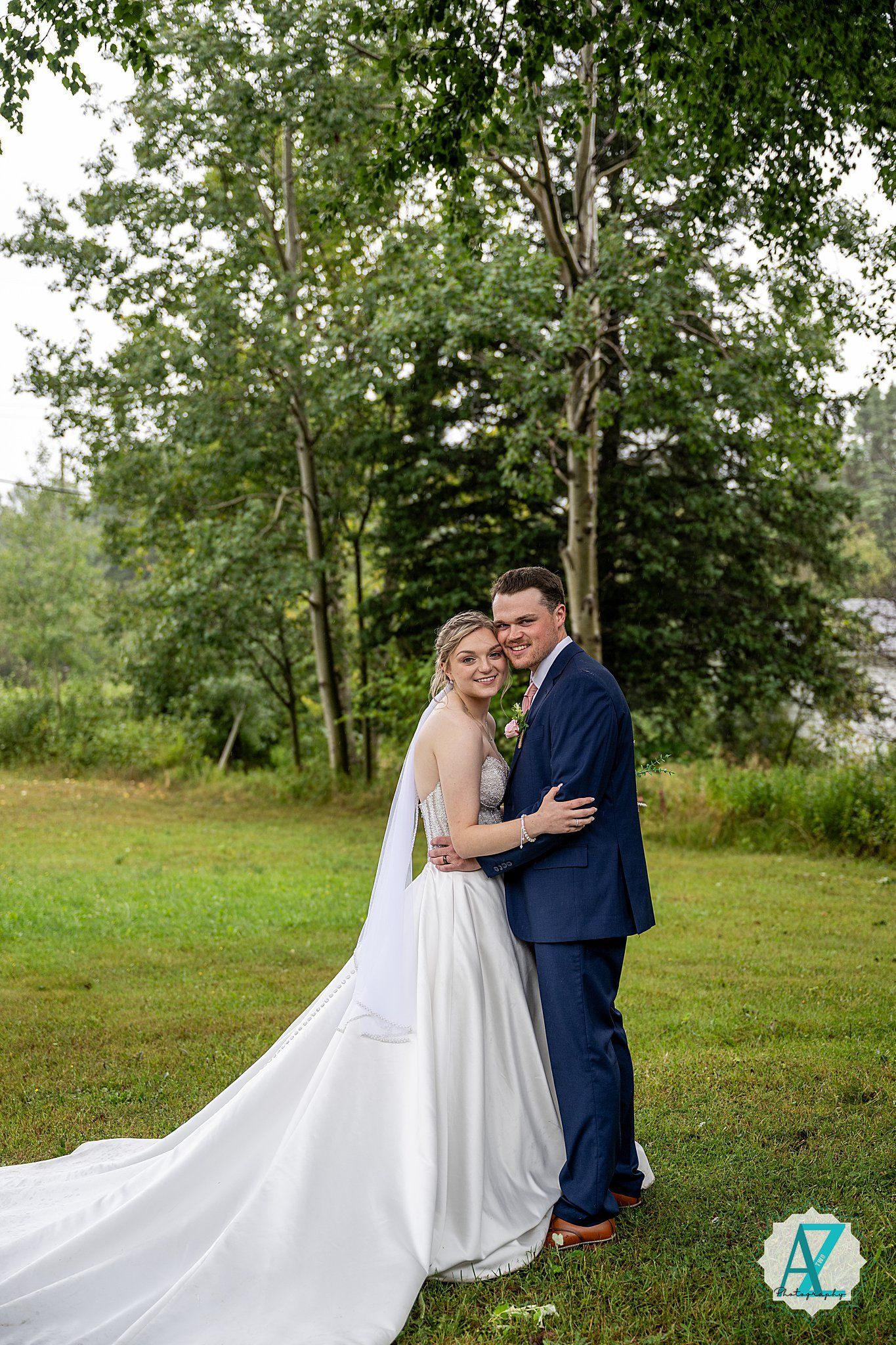 A couple in wedding attire embraces in a lush green outdoor setting with the bride's white dress flowing elegantly.