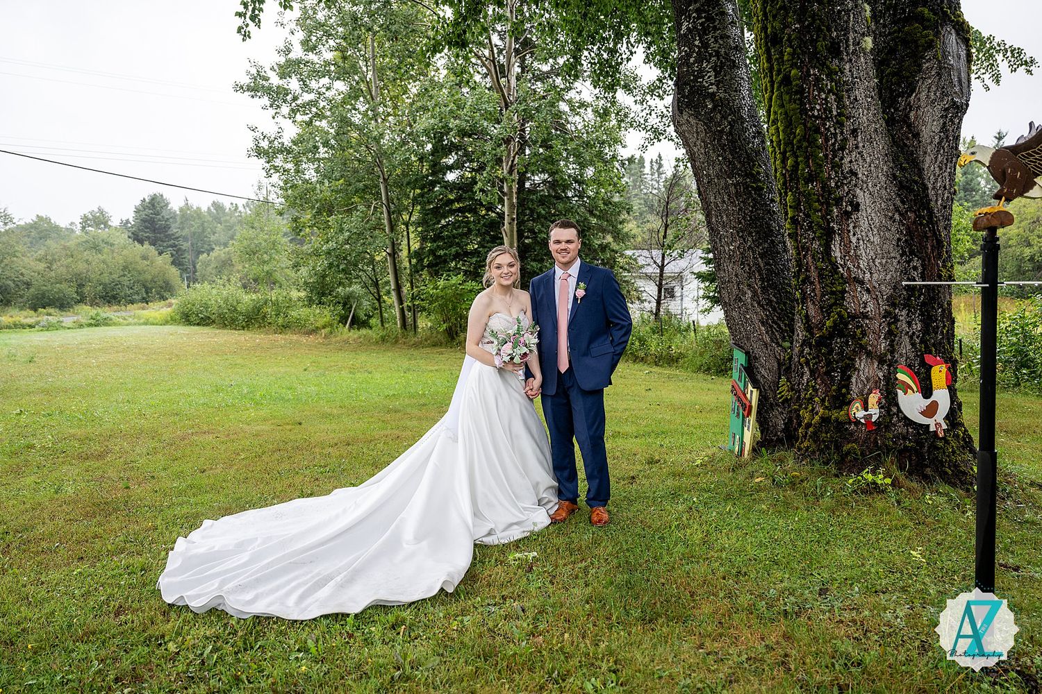 A wedding couple poses under an elegant garden tree canopy in a scenic outdoor setting.