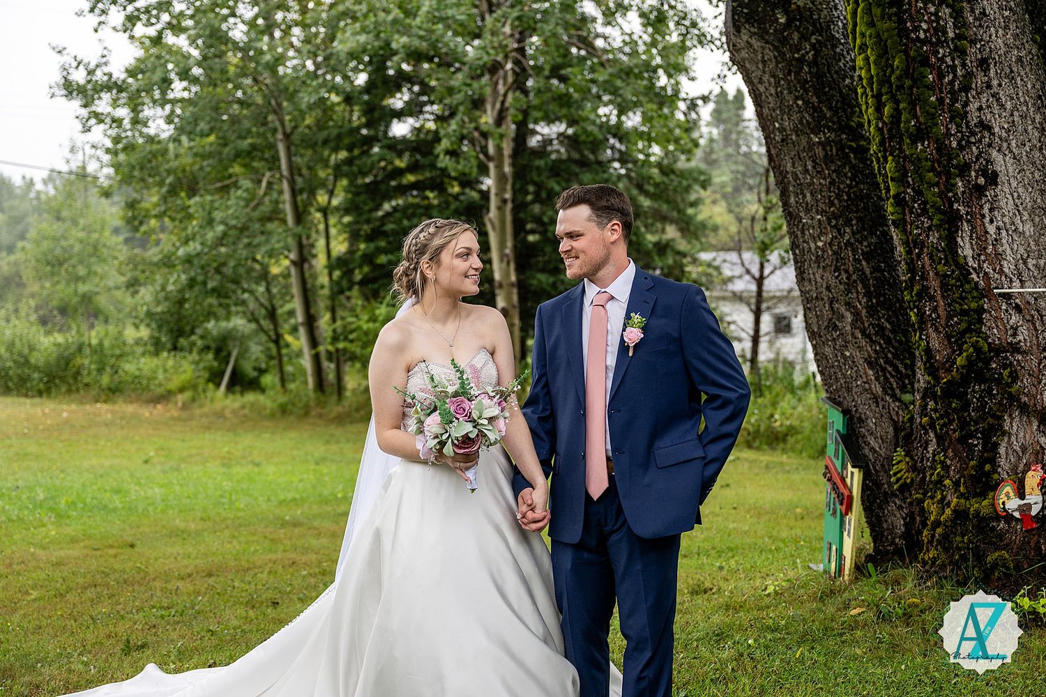 A couple in wedding attire poses on a lush green lawn near tall trees with the bride holding a floral bouquet.